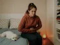 En ung kvinna sitter på sängen med ett **sug i magen**, hennes lilla vita hund vilar bredvid.