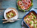 En bit nyttig lasagne på en tallrik bredvid en hel lasagneform och en fräsch tomatsallad med lök.