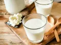 Två glas med krämig kefir står på en träskärbräda. Bredvid syns kefir-kultur och två träskedar. Kefir nyttigt för magen!