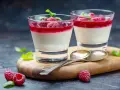 Två glas med sockerfri dessert, panna cotta med hallon och mynta. Serveras på en träskärbräda med skedar och färska hallon.