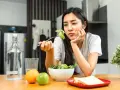 En kvinna sitter vid ett bord med en sallad framför sig, ser ut att tänka "jag kan inte gå ner i vikt". Vattenflaska, frukt och smörgåsar finns också på bordet.