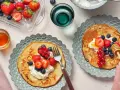 Två tallrikar med bovete pannkakor toppade med grädde och färska bär. En honungsstav droppar honung över den ena pannkakan.