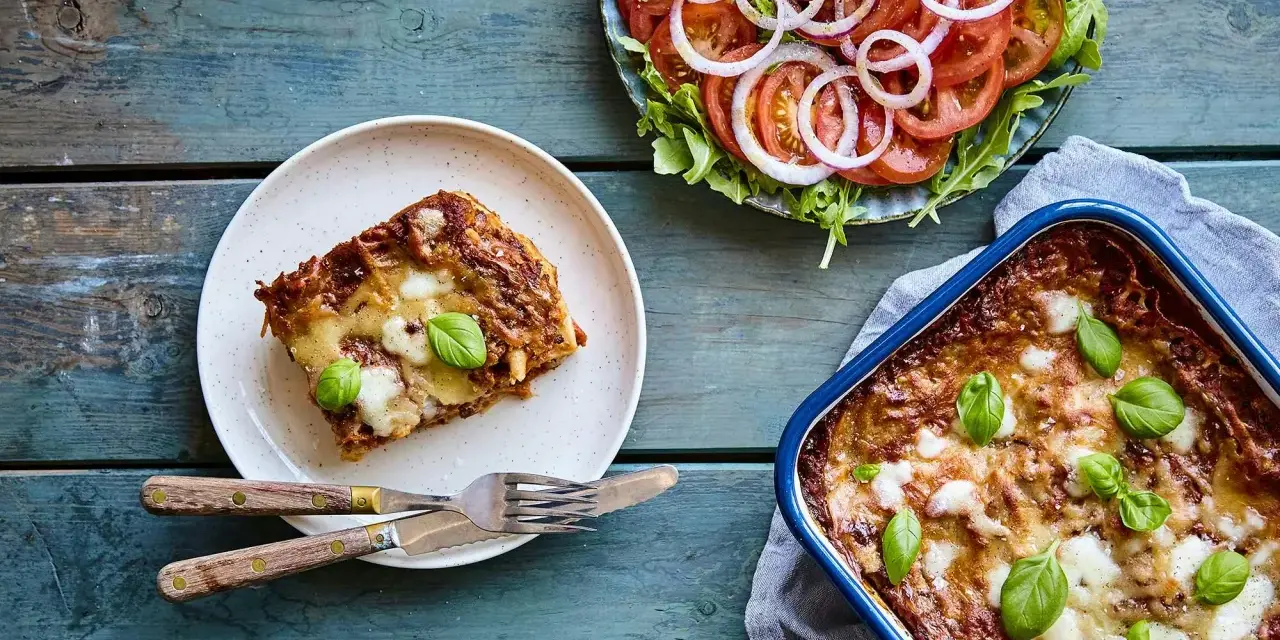 En bit nyttig lasagne på en tallrik bredvid en hel lasagneform och en fräsch tomatsallad med lök.