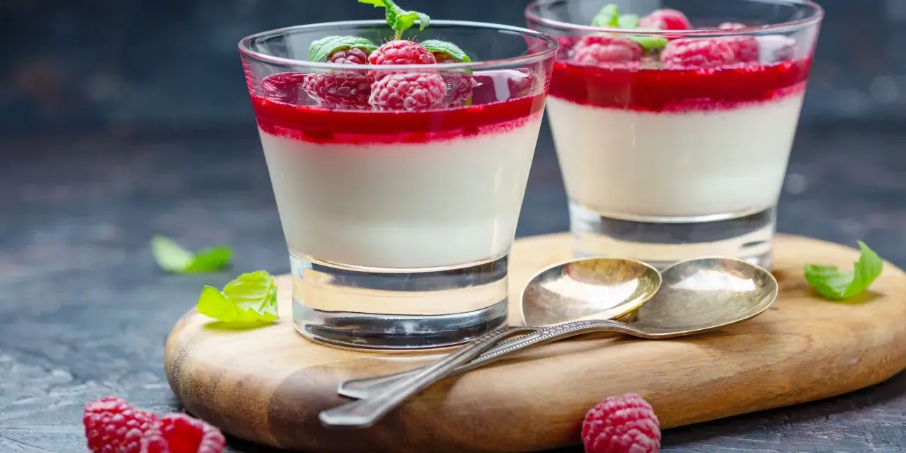 Två glas med sockerfri dessert, panna cotta med hallon och mynta. Serveras på en träskärbräda med skedar och färska hallon.