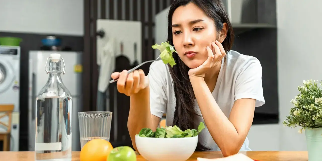 En kvinna sitter vid ett bord med en sallad framför sig, ser ut att tänka "jag kan inte gå ner i vikt". Vattenflaska, frukt och smörgåsar finns också på bordet.