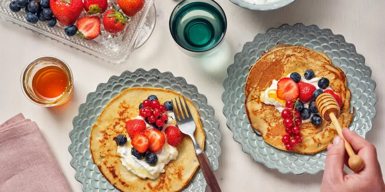 Två tallrikar med bovete pannkakor toppade med grädde och färska bär. En honungsstav droppar honung över den ena pannkakan.