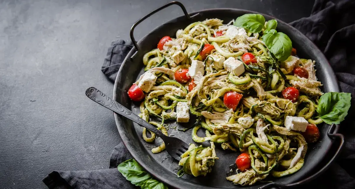 En läcker keto middag med kyckling, zucchinipasta, körsbärstomater och fetaost, garnerad med färsk basilika.