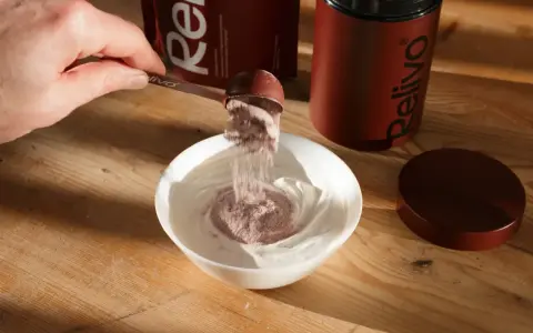 Pulver från en skopa blandas i en skål med yoghurt. En burk och förpackning från Relivo syns i bakgrunden. Perfekt för att dricka mer!