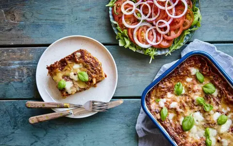 En bit nyttig lasagne på en tallrik bredvid en hel lasagneform och en fräsch tomatsallad med lök.
