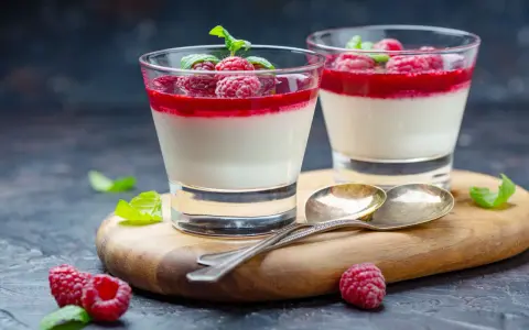 Två glas med sockerfri dessert, panna cotta med hallon och mynta. Serveras på en träskärbräda med skedar och färska hallon.