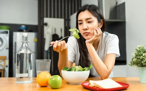 En kvinna sitter vid ett bord med en sallad framför sig, ser ut att tänka "jag kan inte gå ner i vikt". Vattenflaska, frukt och smörgåsar finns också på bordet.