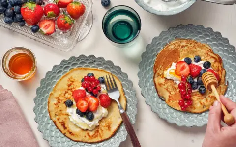 Två tallrikar med bovete pannkakor toppade med grädde och färska bär. En honungsstav droppar honung över den ena pannkakan.