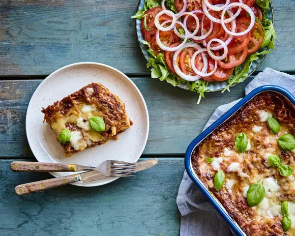 En bit nyttig lasagne på en tallrik bredvid en hel lasagneform och en fräsch tomatsallad med lök.