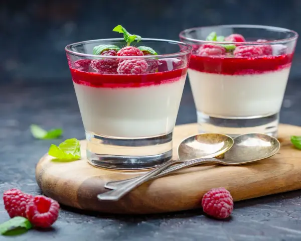Två glas med sockerfri dessert, panna cotta med hallon och mynta. Serveras på en träskärbräda med skedar och färska hallon.