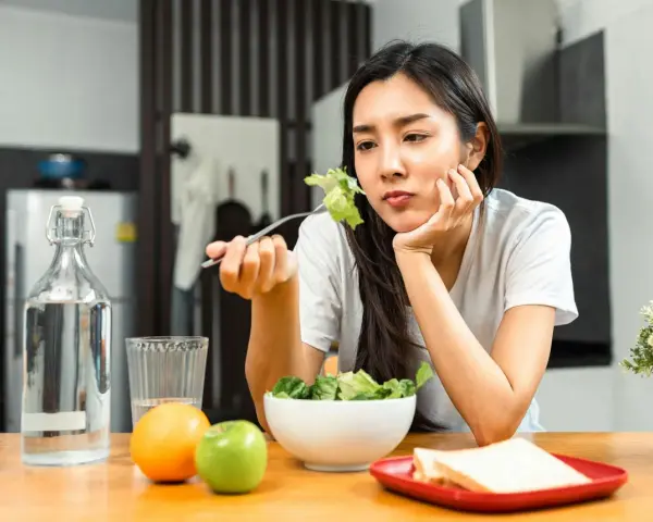 En kvinna sitter vid ett bord med en sallad framför sig, ser ut att tänka "jag kan inte gå ner i vikt". Vattenflaska, frukt och smörgåsar finns också på bordet.