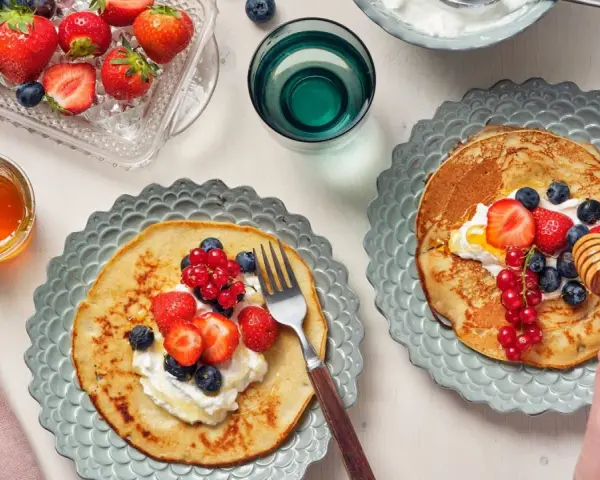 Två tallrikar med bovete pannkakor toppade med grädde och färska bär. En honungsstav droppar honung över den ena pannkakan.