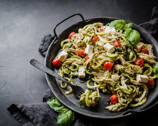 En läcker keto middag med kyckling, zucchinipasta, körsbärstomater och fetaost, garnerad med färsk basilika.