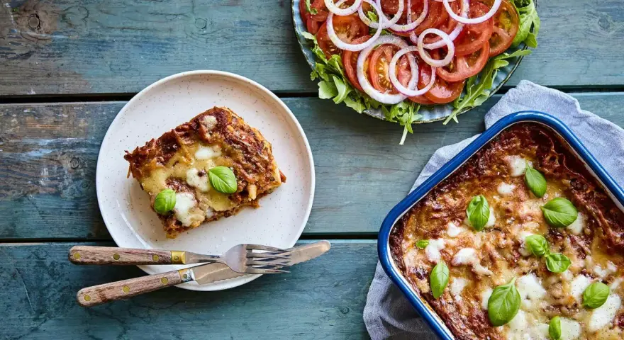En bit nyttig lasagne på en tallrik bredvid en hel lasagneform och en fräsch tomatsallad med lök.