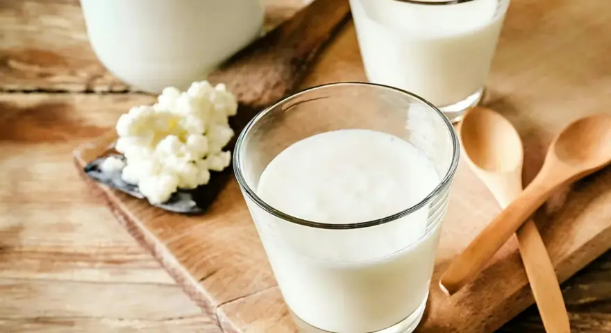 Två glas med krämig kefir står på en träskärbräda. Bredvid syns kefir-kultur och två träskedar. Kefir nyttigt för magen!