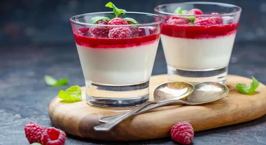 Två glas med sockerfri dessert, panna cotta med hallon och mynta. Serveras på en träskärbräda med skedar och färska hallon.