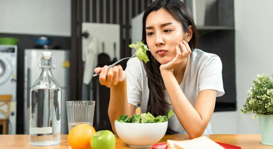 En kvinna sitter vid ett bord med en sallad framför sig, ser ut att tänka "jag kan inte gå ner i vikt". Vattenflaska, frukt och smörgåsar finns också på bordet.