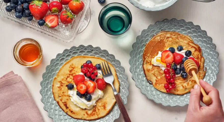 Två tallrikar med bovete pannkakor toppade med grädde och färska bär. En honungsstav droppar honung över den ena pannkakan.