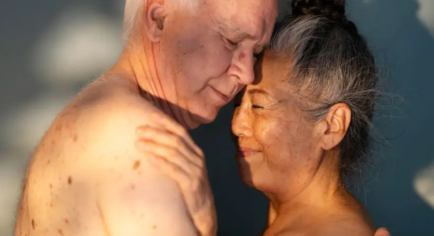 Ett äldre par omfamnar varandra ömt, deras ansikten nära varandra. Ljuset faller mjukt över dem och framhäver deras hud. Kärlek och närhet lyser igenom, en påminnelse om att bra sex kan finnas i alla åldrar.