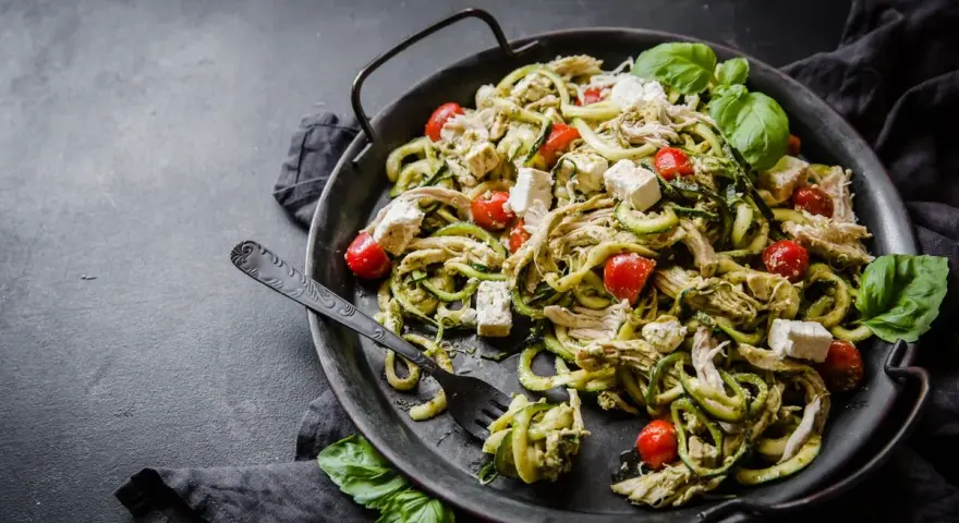En läcker keto middag med kyckling, zucchinipasta, körsbärstomater och fetaost, garnerad med färsk basilika.