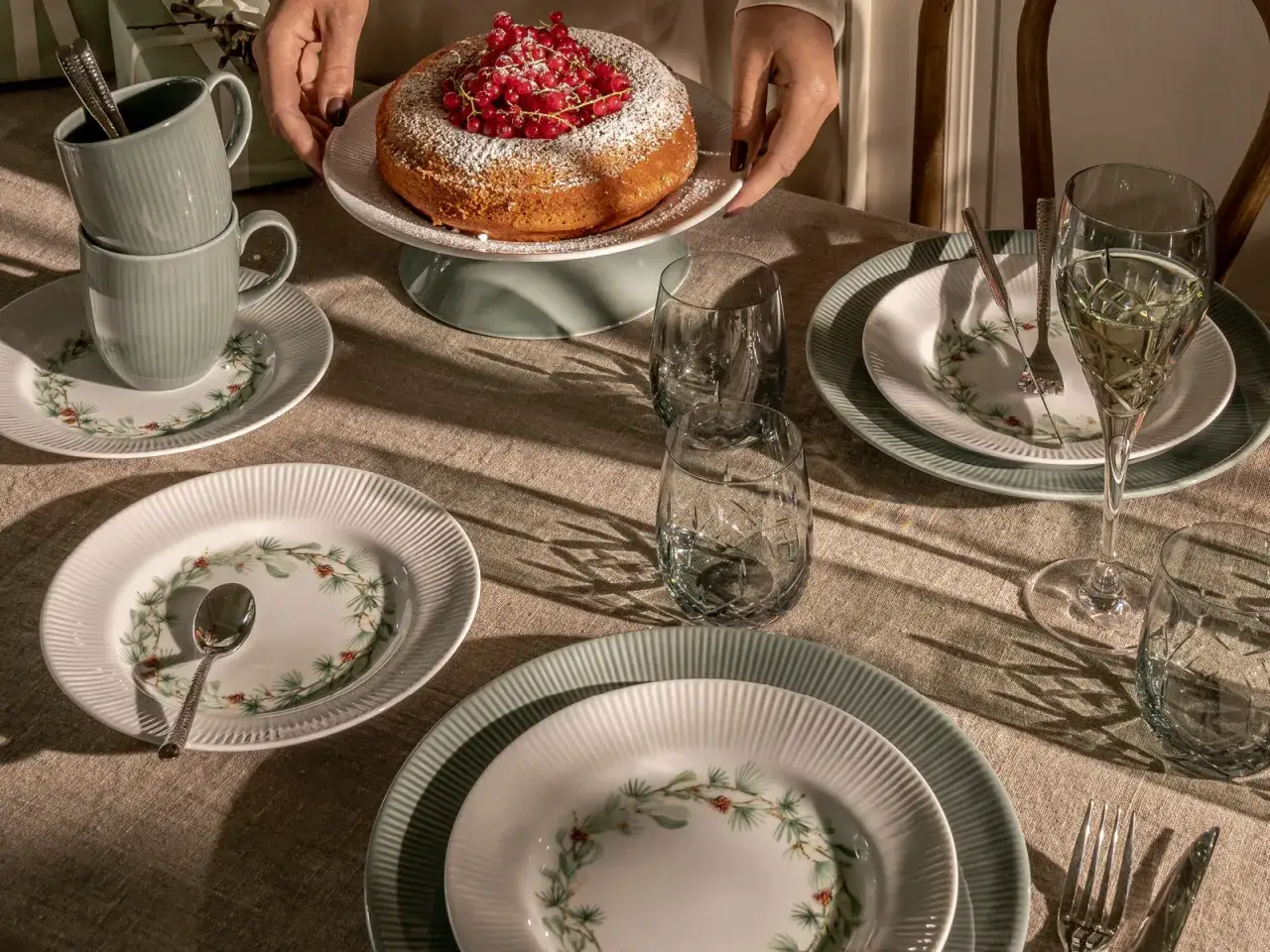 Vacker dukning med en sockerkaka toppad med vinbär, som en hyllning till nordisk kost. Tallrikar med granris och muggar i en lugn färgskala.