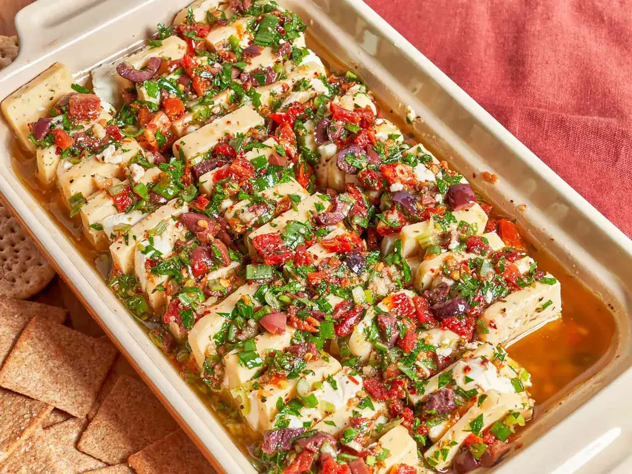 Baked feta cheese with sun-dried tomatoes, olives, herbs, and oil in a baking dish, with crackers on the side.