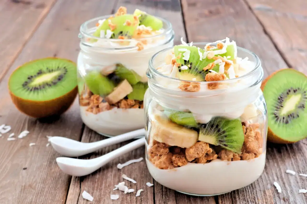 Två glas med en nyttig frukost av yoghurt, granola, kiwi och banan. Serveras på en träbakgrund med kokosflingor.