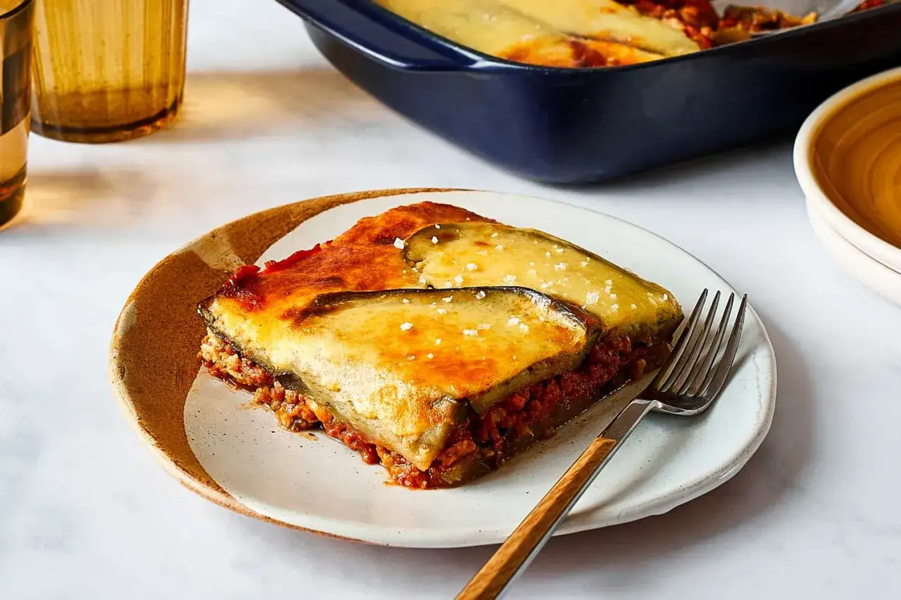 En bit lchf moussaka med aubergine och k&ouml;ttf&auml;rss&aring;s, toppad med ost och flingsalt. Serveras p&aring; en tallrik med en gaffel.
