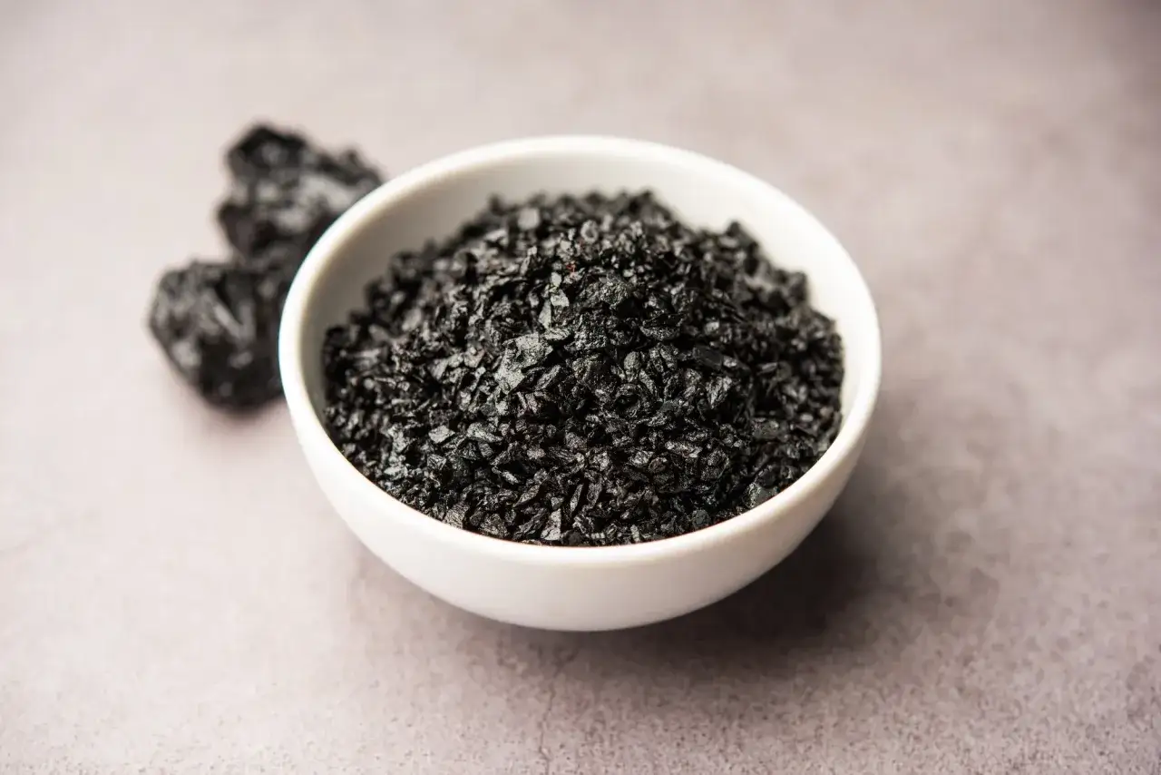 En vit sk&aring;l fylld med svart shilajit-pulver. N&aring;gra klumpar av shilajit syns till v&auml;nster. Viktigt att k&auml;nna till shilajit biverkningar.