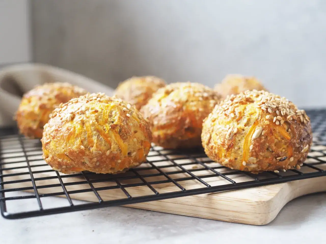 Nybakade glutenfria morotsfrallor med sesamfr&ouml;n och solrosfr&ouml;n p&aring; ett svart galler.